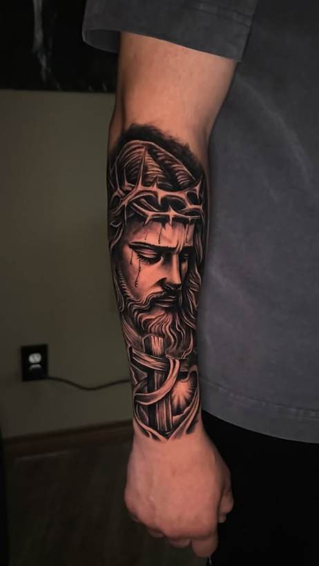 Black and grey portrait forearm piece with a cross. Tattoo by a Thorn and Oath artist.